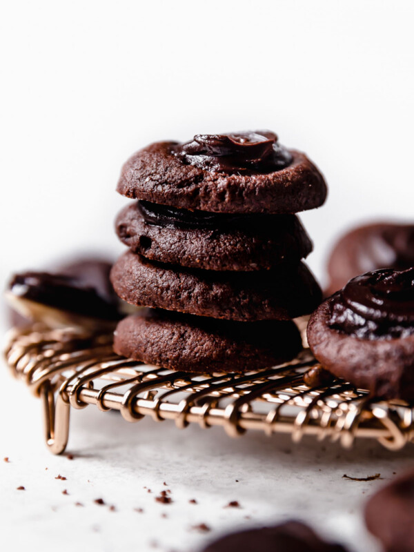 A stack of 4 double chocolate thumbprint cookies placed on a copper wire cooling rack. The stack is surrounded by additional cookies & a spoonful of fudgy ganache.
