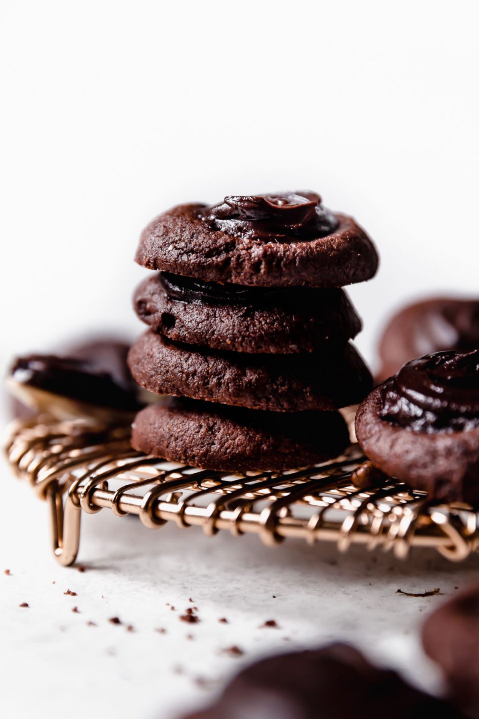 A stack of 4 double chocolate thumbprint cookies placed on a copper wire cooling rack. The stack is surrounded by additional cookies & a spoonful of fudgy ganache.