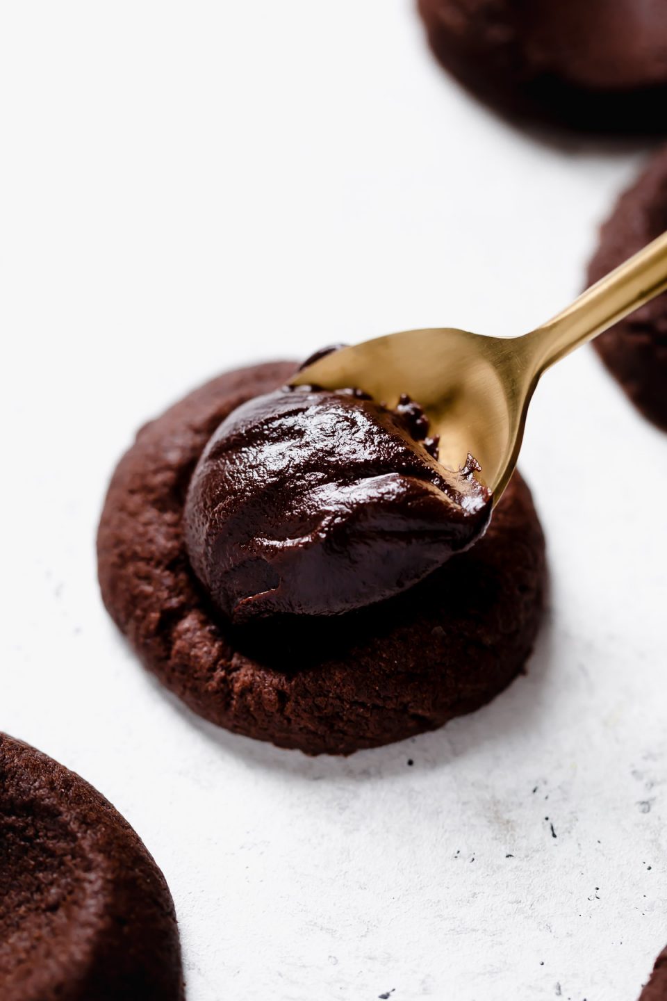 Gold spoon adding chocolate ganache to a baked chocolate thumbprint cookie.