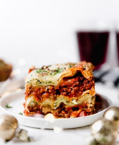 Christmas Eve Lasagna on a small white plate, atop a white surface surrounded by small Christmas ornaments & a glass of red wine.