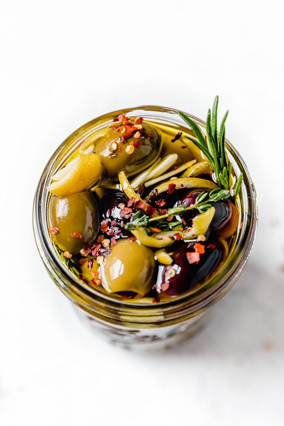 Marinated olives in a jar, topped with chili flake, lemon rind & fresh rosemary.