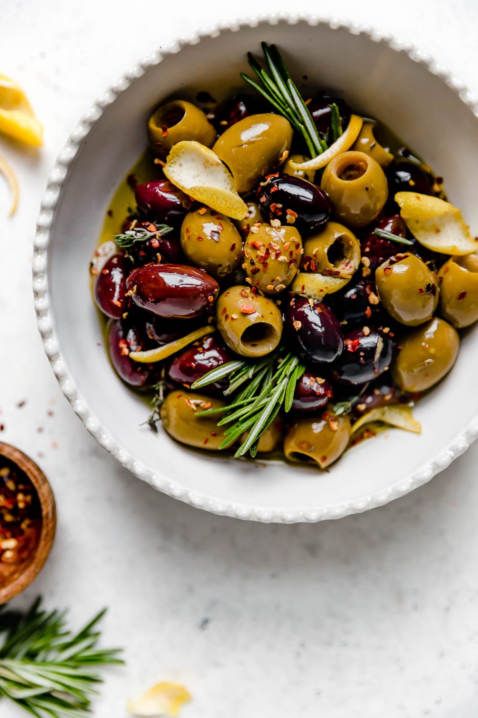 Homemade marinated olives served in a white dish with scalloped edges. The bowl is atop a white surface, surrounded by fresh rosemary, lemon peel, & a small wooden bowl with red chili flakes.
