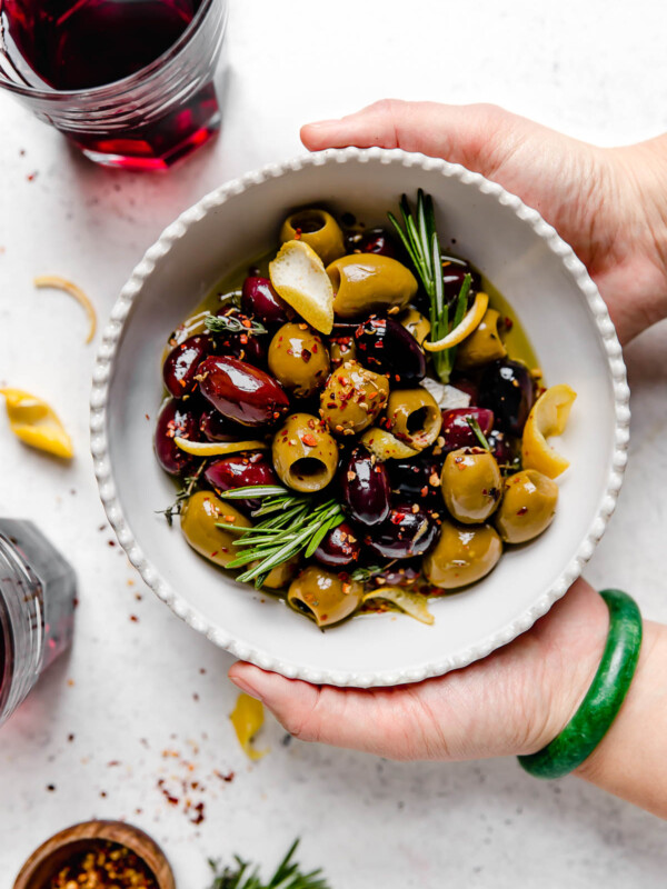 Homemade marinated olives served in a white dish with scalloped edges. The bowl is atop a white surface, surrounded by fresh rosemary, lemon peel, a small wooden bowl with red chili flakes, & a few glasses of red wine. A woman's hands are reaching into the photo, holding the bowl of olives.