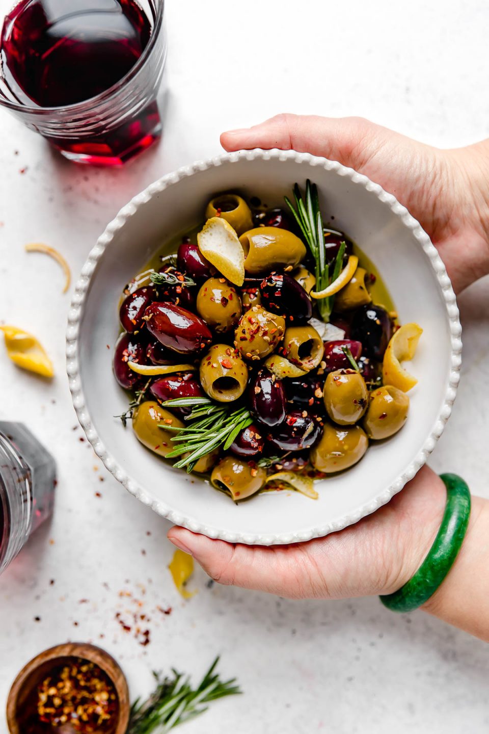 Homemade marinated olives served in a white dish with scalloped edges. The bowl is atop a white surface, surrounded by fresh rosemary, lemon peel, a small wooden bowl with red chili flakes, & a few glasses of red wine. A woman's hands are reaching into the photo, holding the bowl of olives.
