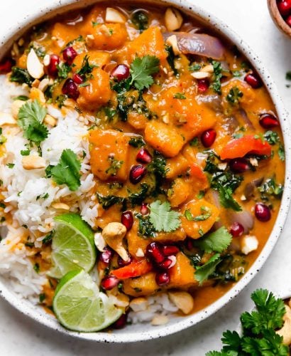 Butternut Squash Red Curry in a speckled white bowl, with white rice, chopped cilantro, cashews, & pomegranate arils.