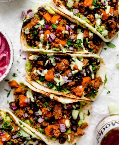 Chorizo Sweet Potato Tacos arranged on a white surface. The tacos are topped with a drizzle of homemade cilantro lime crema. Surrounding the tacos are small bowls of taco toppings (pickled red onions, guacamole, crema, etc.) & a bottle of Modelo beer.