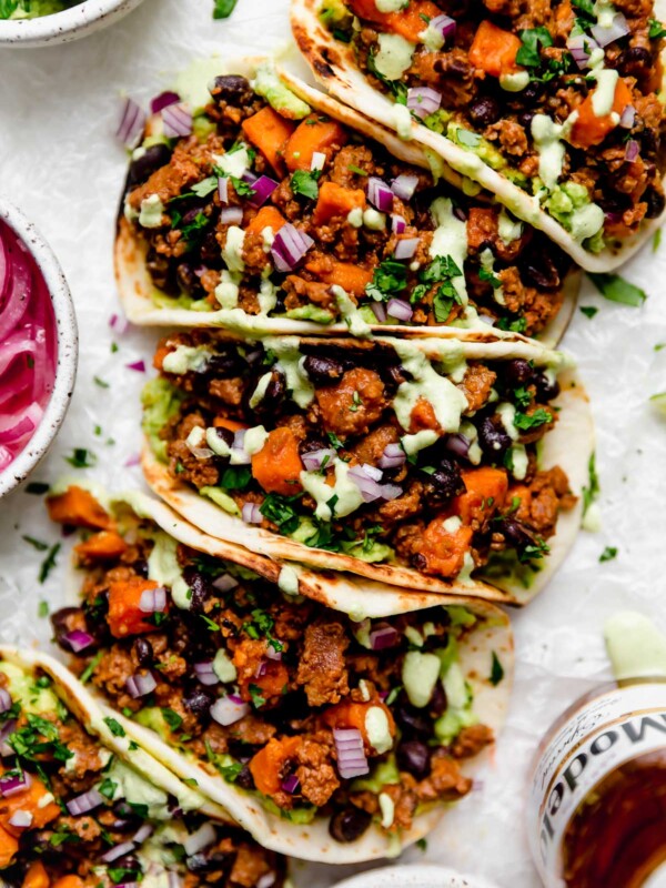 Chorizo Sweet Potato Tacos arranged on a white surface. The tacos are topped with a drizzle of homemade cilantro lime crema. Surrounding the tacos are small bowls of taco toppings (pickled red onions, guacamole, crema, etc.) & a bottle of Modelo beer.