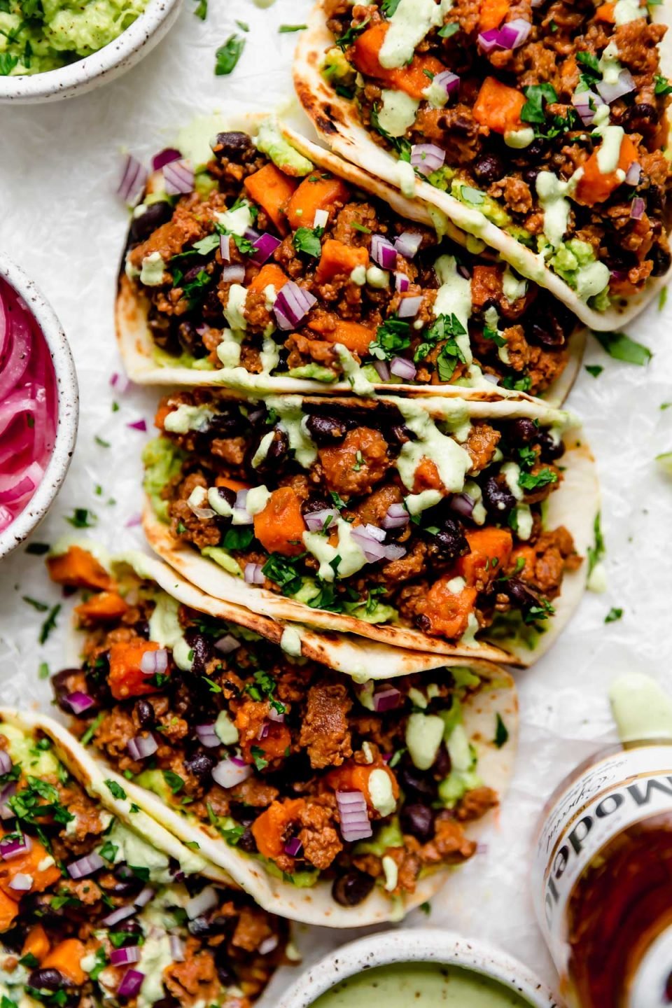 Chorizo Sweet Potato Tacos arranged on a white surface. The tacos are topped with a drizzle of homemade cilantro lime crema. Surrounding the tacos are small bowls of taco toppings (pickled red onions, guacamole, crema, etc.) & a bottle of Modelo beer.