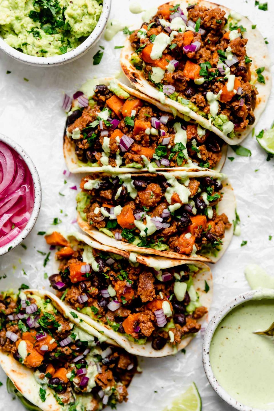Chorizo Sweet Potato Tacos arranged on a white surface. The tacos are topped with a drizzle of homemade cilantro lime crema. Surrounding the tacos are small bowls of taco toppings (pickled red onions, guacamole, crema, etc.).