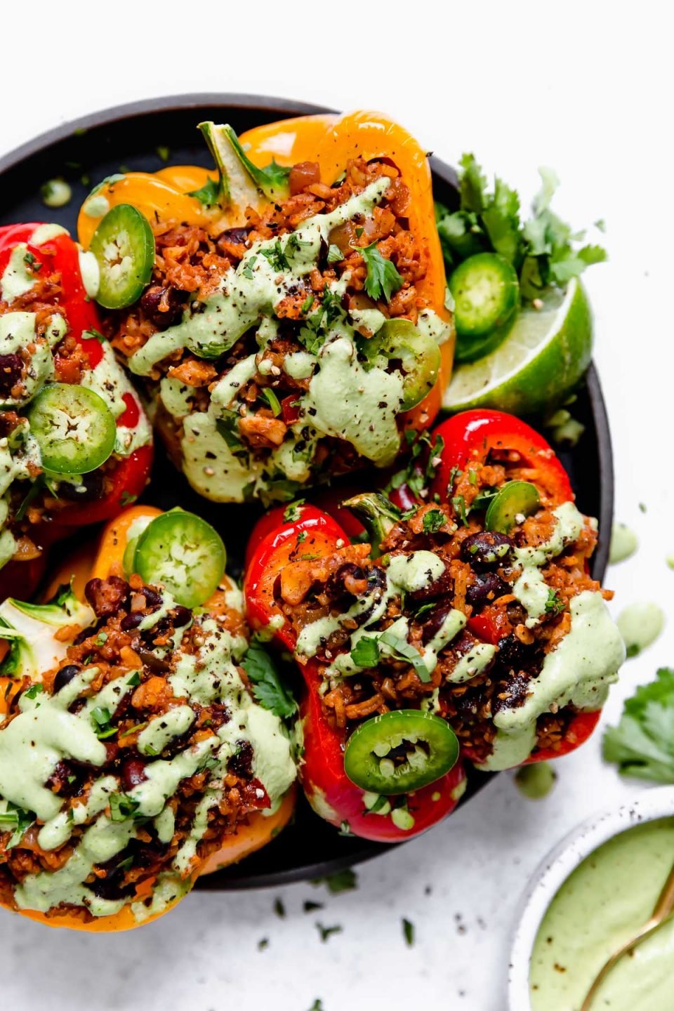 4 Vegan Stuffed Peppers on a small black plate, topped with cilantro lime cashew crema, sliced jalapenos, lime wedges & cilantro.