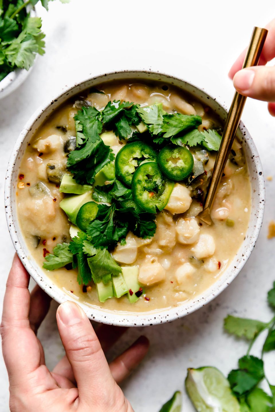 Vegan white chili served in a speckled ceramic bowl, topped with sliced jalapeno, cilantro, & diced avocado. A woman's hands are reaching in, with a large gold spoon, to take a bite of the chili.