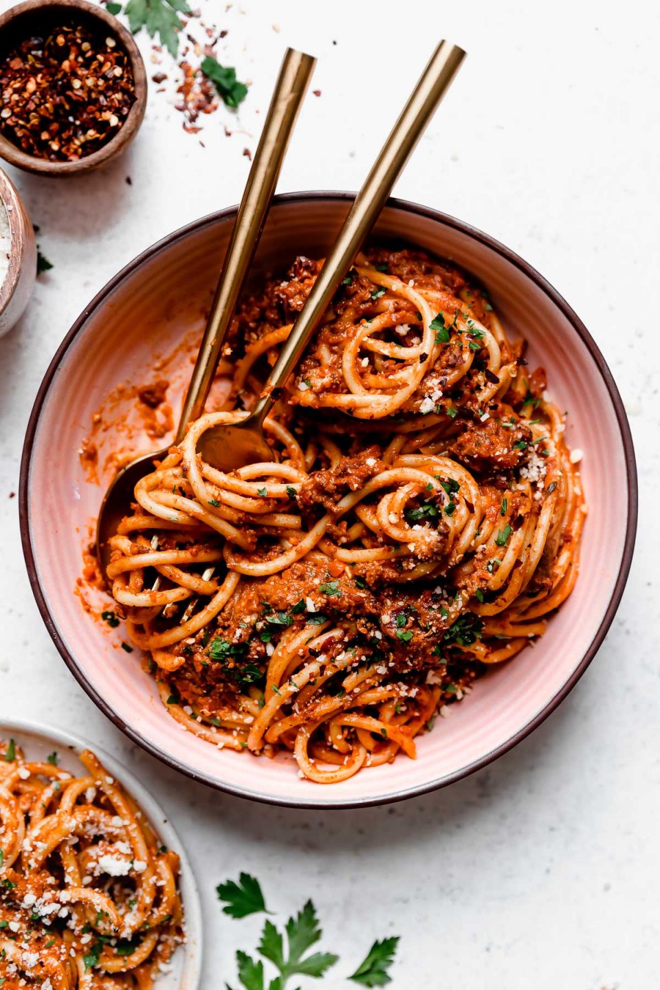 Bolognese sauce tossed into bucatini noodles, served in a pink pasta bowl with gold flatware. The pasta bolognese is topped with grated cheese, chopped parsley, & crushed red pepper flakes.