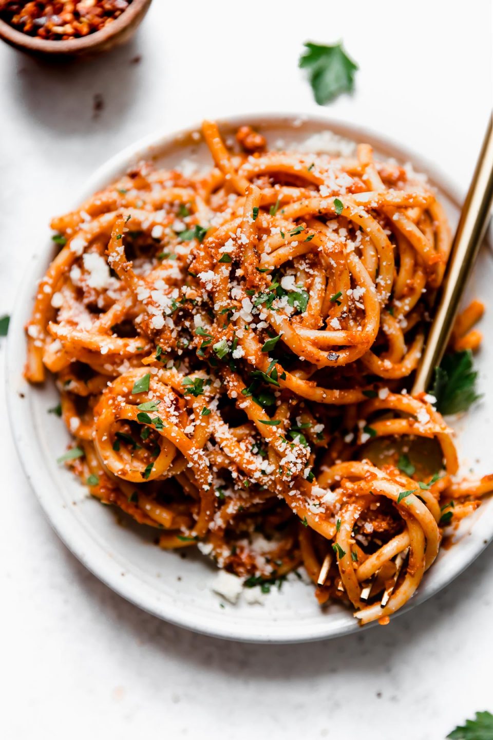 Bolognese sauce tossed into bucatini noodles, served on a small white plate with gold flatware. The pasta bolognese is topped with grated cheese, chopped parsley, & crushed red pepper flakes.