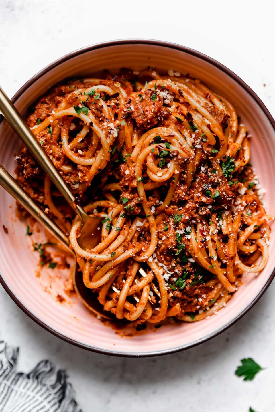 Bolognese sauce tossed into bucatini noodles, served in a pink pasta bowl with gold flatware. The pasta bolognese is topped with grated cheese, chopped parsley, & crushed red pepper flakes.