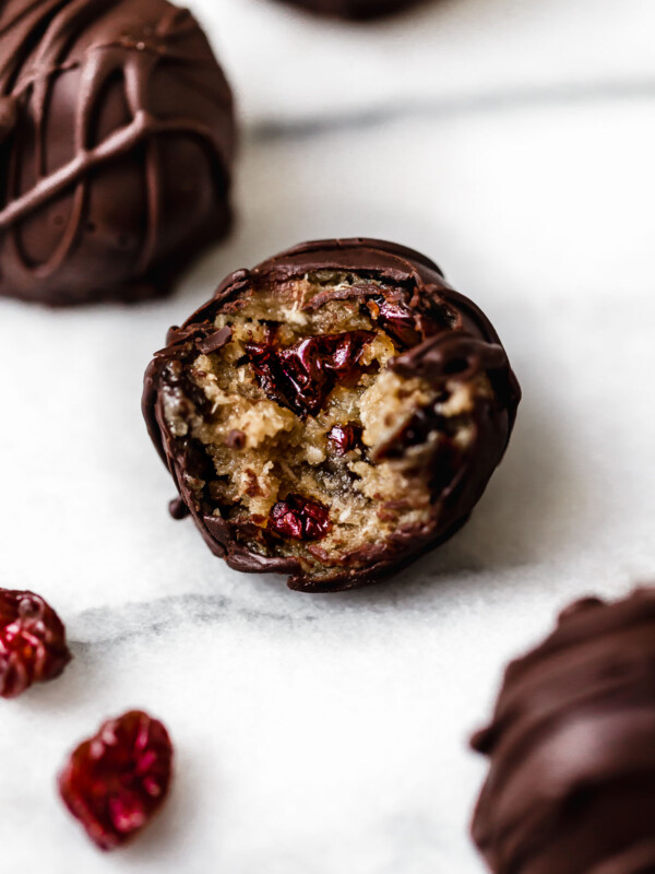 Chocolate-covered vegan cookie dough bite sitting on a white surface. A bite has been taken out of the ball, revealing an inside studded with ruby-red Montmorency tart cherries.