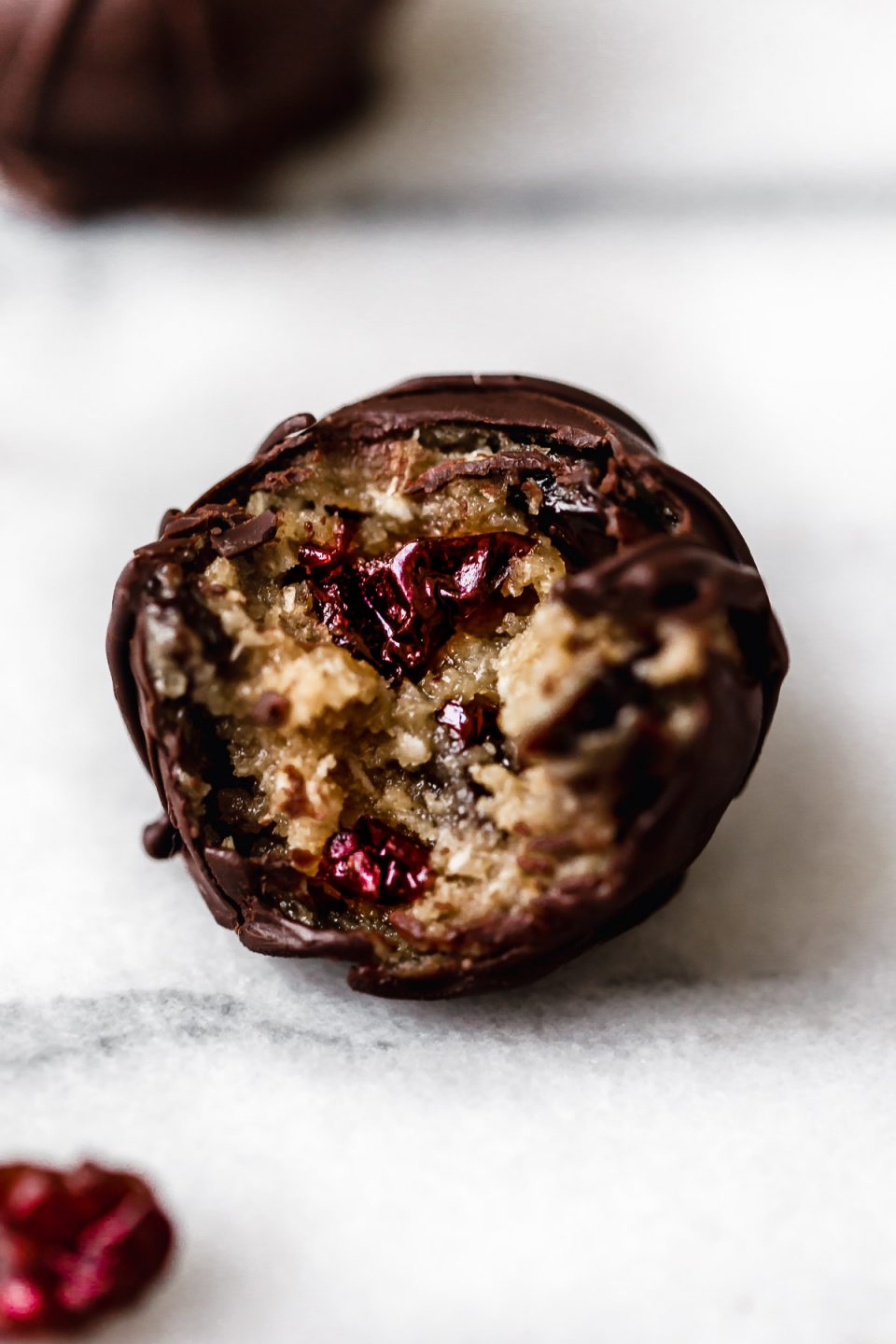 Chocolate-covered vegan cookie dough bite sitting on a white surface. A bite has been taken out of the ball, revealing an inside studded with ruby-red Montmorency tart cherries.