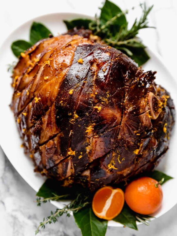 Citrus & Maple Glazed ham on a white serving platter, with fresh bay leaves, fresh rosemary, fresh thyme, & oranges.