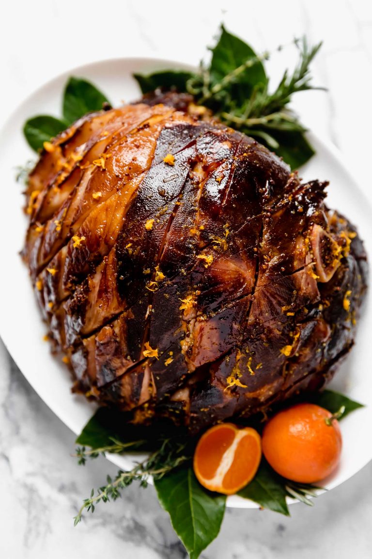 Citrus & Maple Glazed ham on a white serving platter, with fresh bay leaves, fresh rosemary, fresh thyme, & oranges.