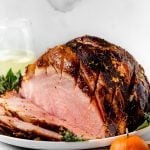 Citrus & Maple Glazed ham (shown partially sliced) on a white serving platter, with a glass of white wine, fresh bay leaves, fresh rosemary, fresh thyme, & oranges.