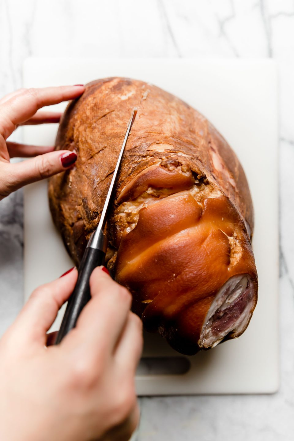 A woman's hands shown using a paring knife to score the surface of a ham shank. 