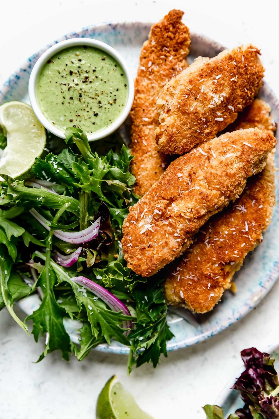 Crispy coconut chicken tenders on a blue-speckled plate with salad greens and cilantro-lime cashew crema for dipping.
