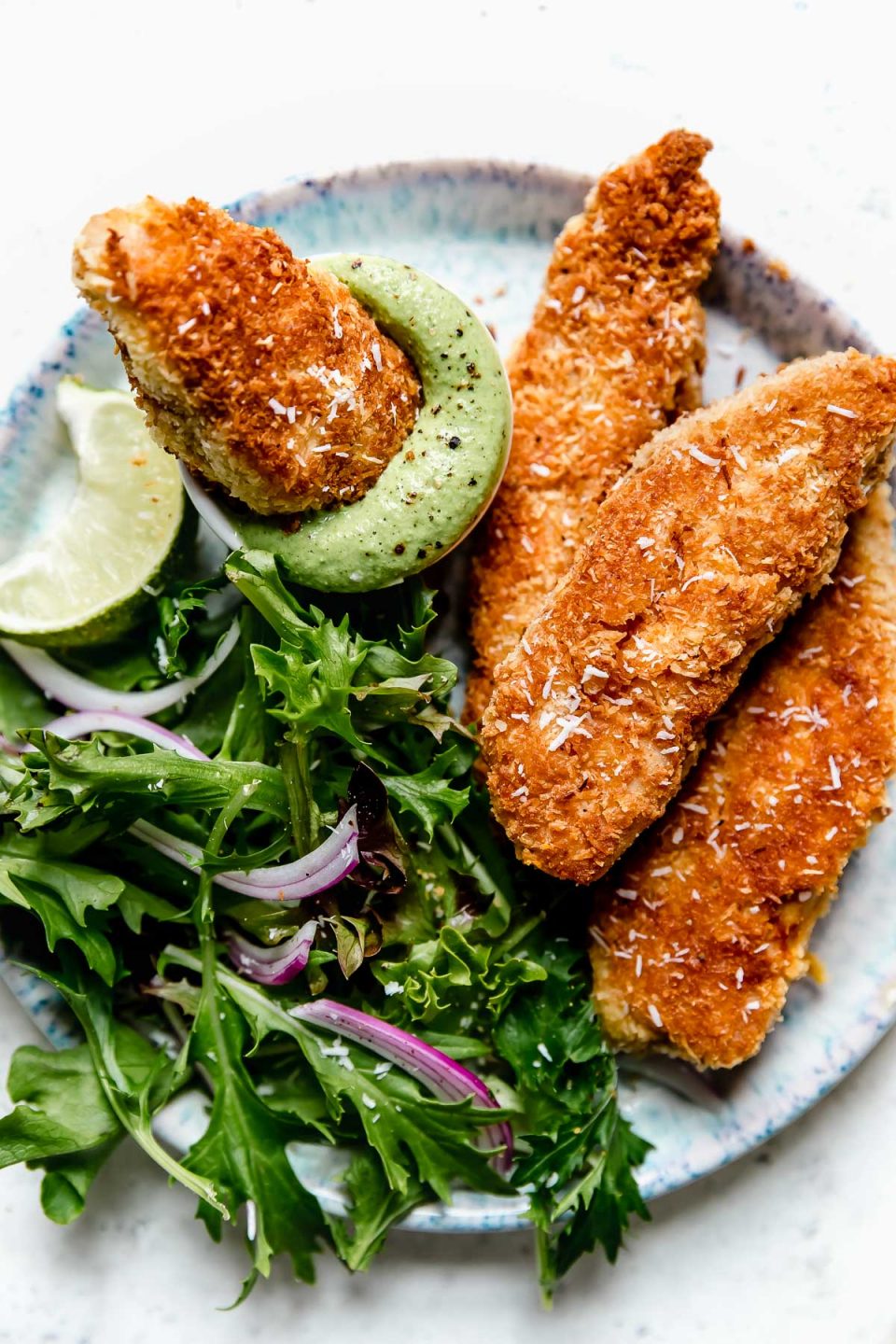 Crispy coconut chicken tenders on a blue-speckled plate with salad greens and cilantro-lime cashew crema for dipping.