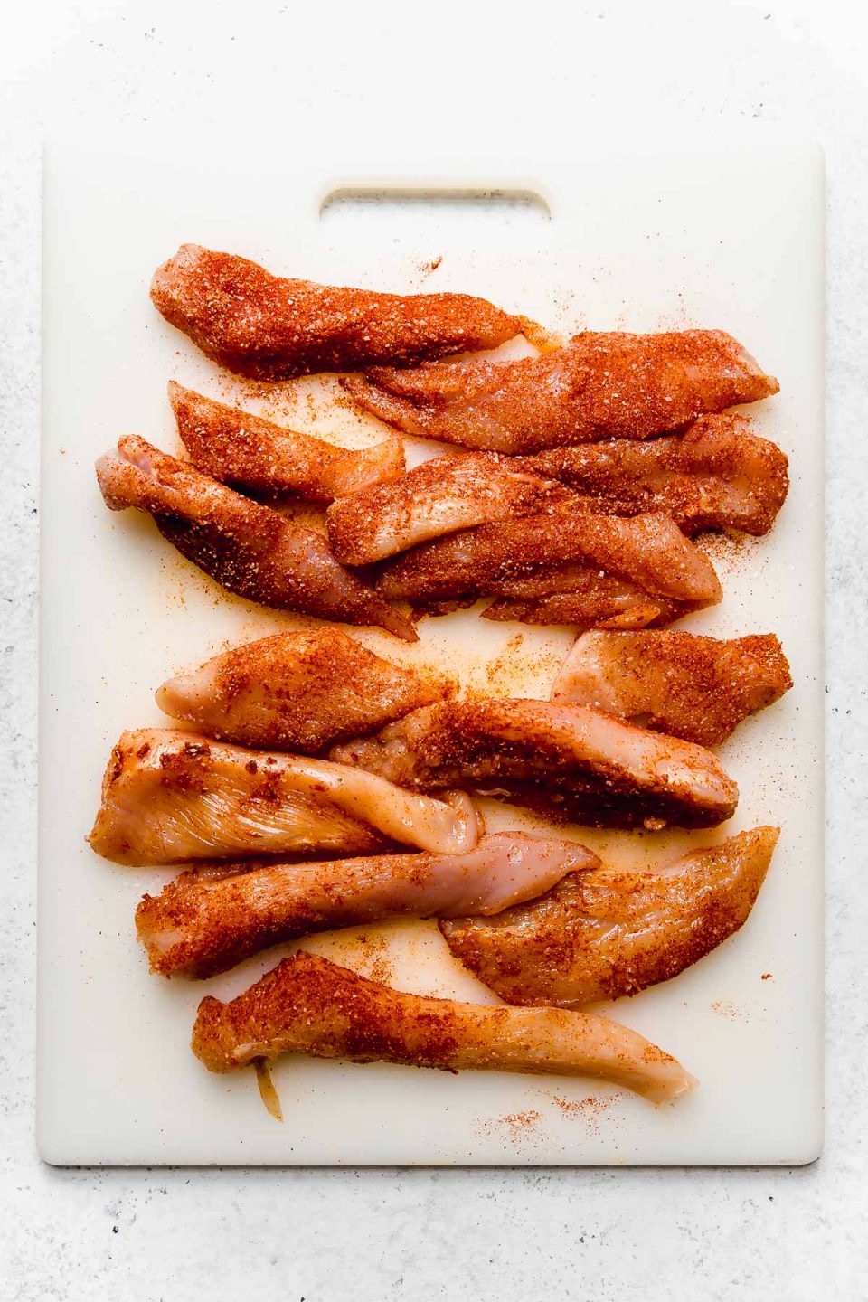 Chicken breast strips, seasoned in paprika, on a white cutting board.