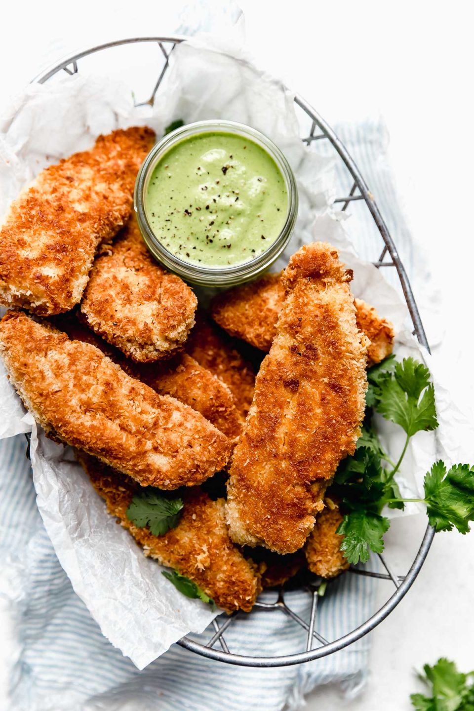 Golden brown coconut chicken tenders in a paper-lined wire basket with cilantro-lime cashew crema. The basket sits atop a blue & white striped linen.