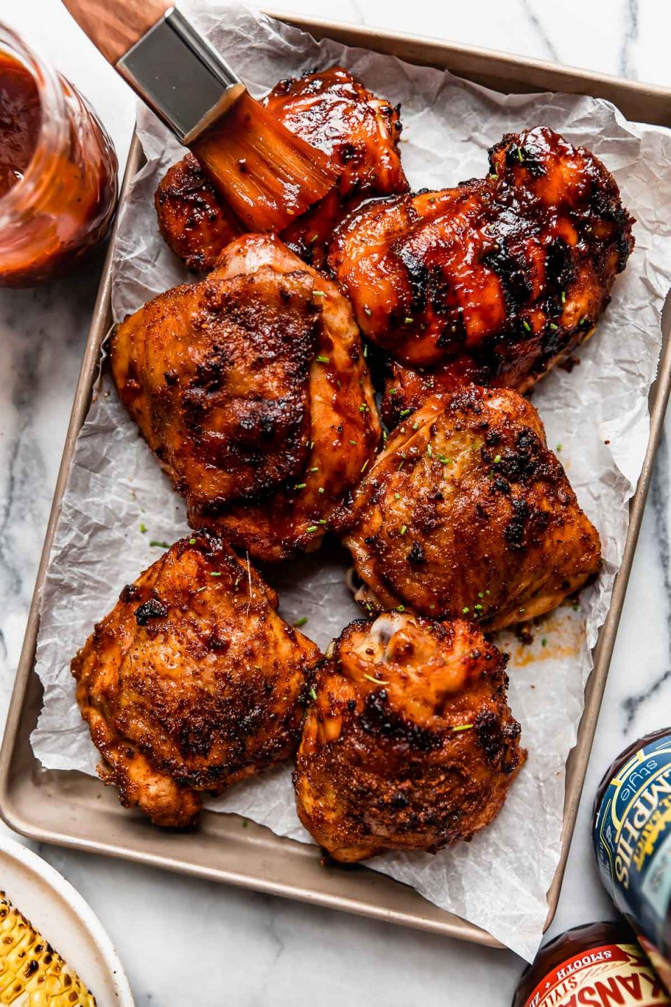 6 grilled BBQ chicken thighs on a small baking sheet. Some of the chicken pieces have crispy skin. Others are slathered in BBQ sauce. A pastry brush is reaching in from outside of the frame, brushing even more BBQ sauce on the barbecue chicken. 
