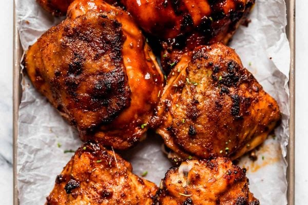6 grilled BBQ chicken thighs on a small baking sheet. Some of the chicken pieces are slathered in barbecue sauce, while others have crispy skin.