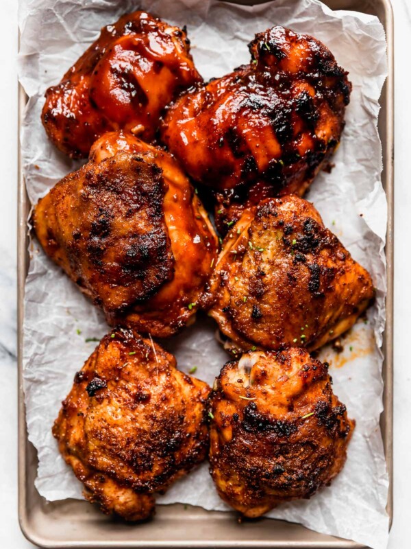6 grilled BBQ chicken thighs on a small baking sheet. Some of the chicken pieces are slathered in barbecue sauce, while others have crispy skin.