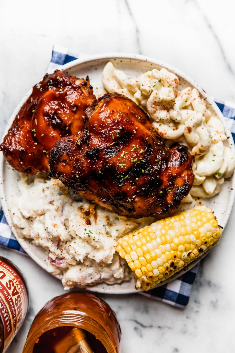 Grilled BBQ chicken on a plate with macaroni salad, potato salad, & corn on the cob. Next to the plate is a bottle of barbecue sauce from ALDI.