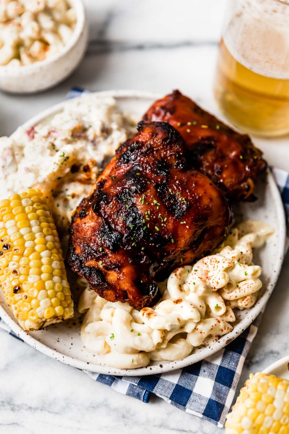 Grilled BBQ chicken on a plate with macaroni salad, potato salad, & corn on the cob. The plate is surrounded by more side dishes, extra BBQ sauce & a glass of beer.