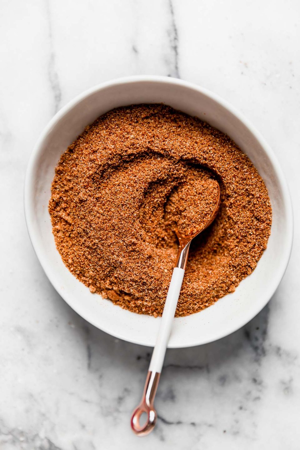 The BEST dry rub for grilling in a small white bowl, set on a white marble surface. There is a small white spoon in the bowl, holding some of the barbecue dry rub.