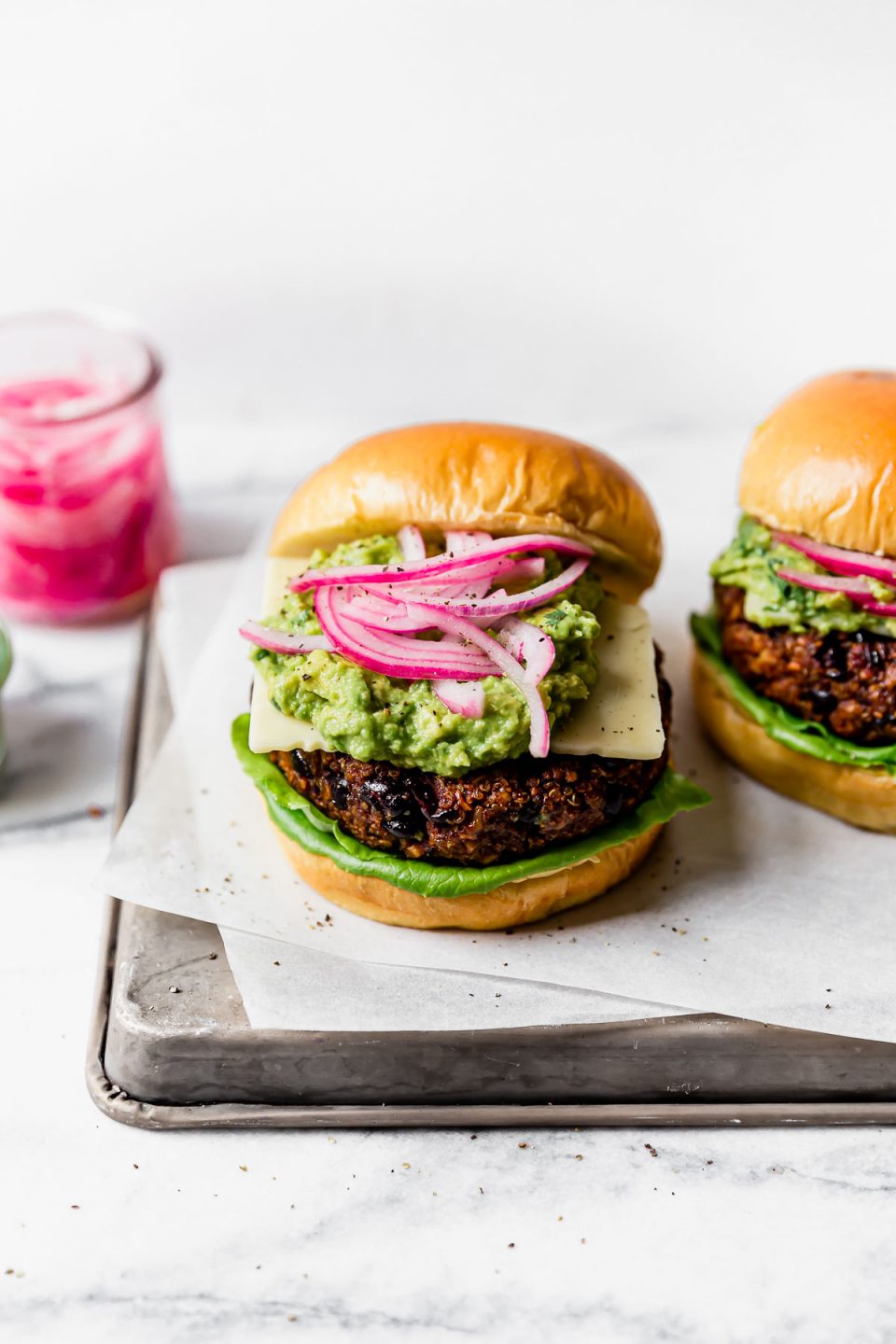 Grilled Black Bean Burgers on brioche buns, topped with lettuce, cheese, guacamole & pickled red onion. The burgers are placed on a small metal tray, with jars of guacamole & pickled red onions in the background.