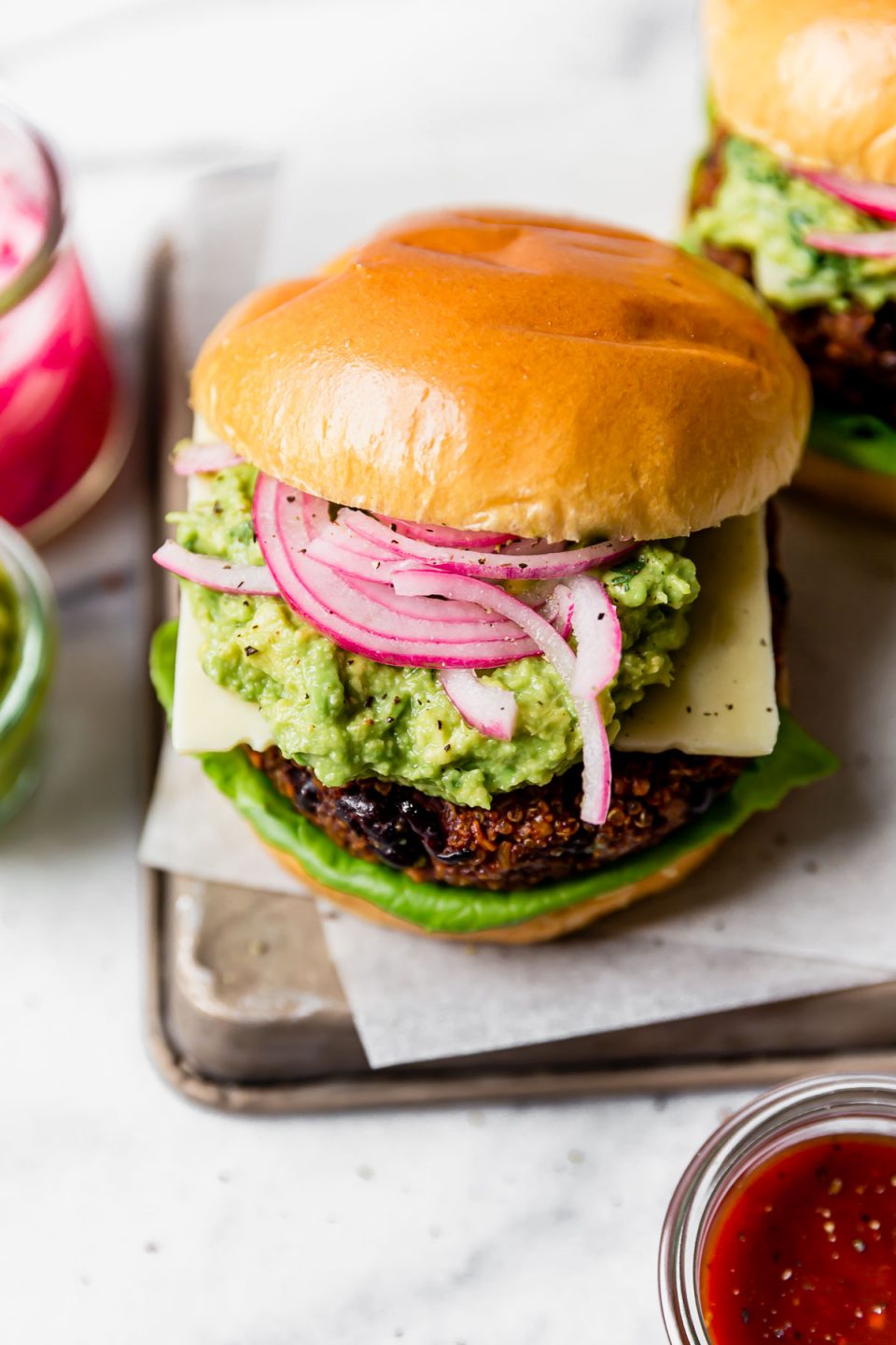Overhead view of grilled Black Bean Burgers on brioche buns, topped with lettuce, cheese, guacamole & pickled red onion. The burgers are placed on a small metal tray, with jars of guacamole & pickled red onions in the background.