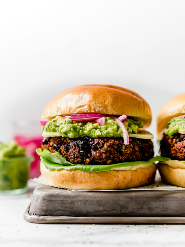 Grilled Black Bean Burgers on brioche buns, topped with lettuce, cheese, guacamole & pickled red onion. The burgers are placed on a small metal tray, with jars of guacamole & pickled red onions in the background.
