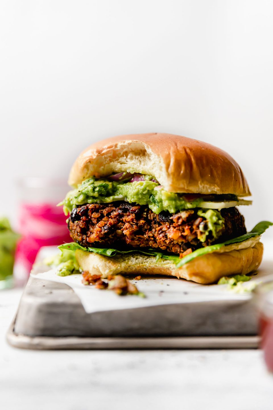 Grilled Black Bean Burgers with a large bite taken out of it. The burger is placed on a small metal tray, with jars of guacamole & pickled red onions in the background.