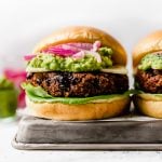 Grilled Black Bean Burgers on brioche buns, topped with lettuce, cheese, guacamole & pickled red onion. The burgers are placed on a small metal tray, with jars of guacamole & pickled red onions in the background.