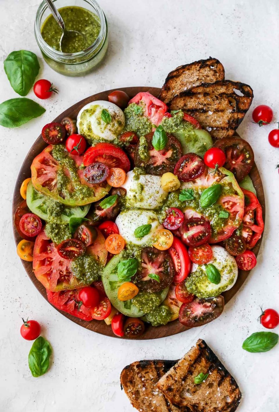 Heirloom tomato burrata salad arranged on a large wooden serving board, topped with vibrant green fresh pesto sauce. The board is surrounded by a few slices of grilled bread, fresh basil leaves, & a jar of pesto sauce atop a neutral background.
