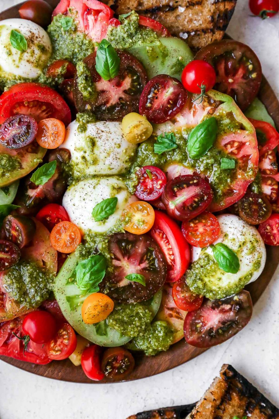 Heirloom tomato burrata salad arranged on a large wooden serving board, topped with vibrant green fresh pesto sauce. The board is surrounded by a few slices of grilled bread.