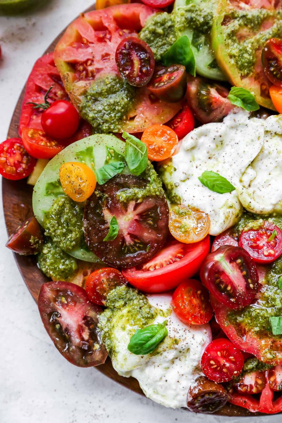 Heirloom tomato burrata salad arranged on a large wooden serving board, topped with vibrant green fresh pesto sauce. The board is surrounded by a few slices of grilled bread & some red cherry tomatoes.