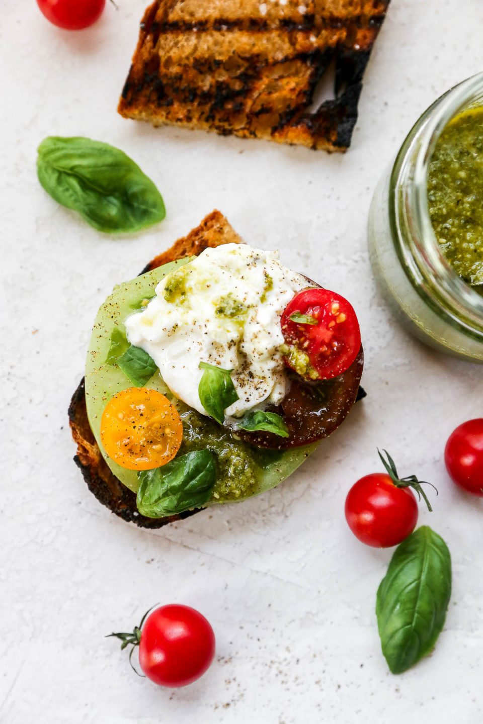 Burrata Caprese Salad served on a piece of grilled bread. The bread sits atop a neutral background, surrounded by whole cherry tomatoes, fresh bail leaves & a jar of basil pesto sauce.