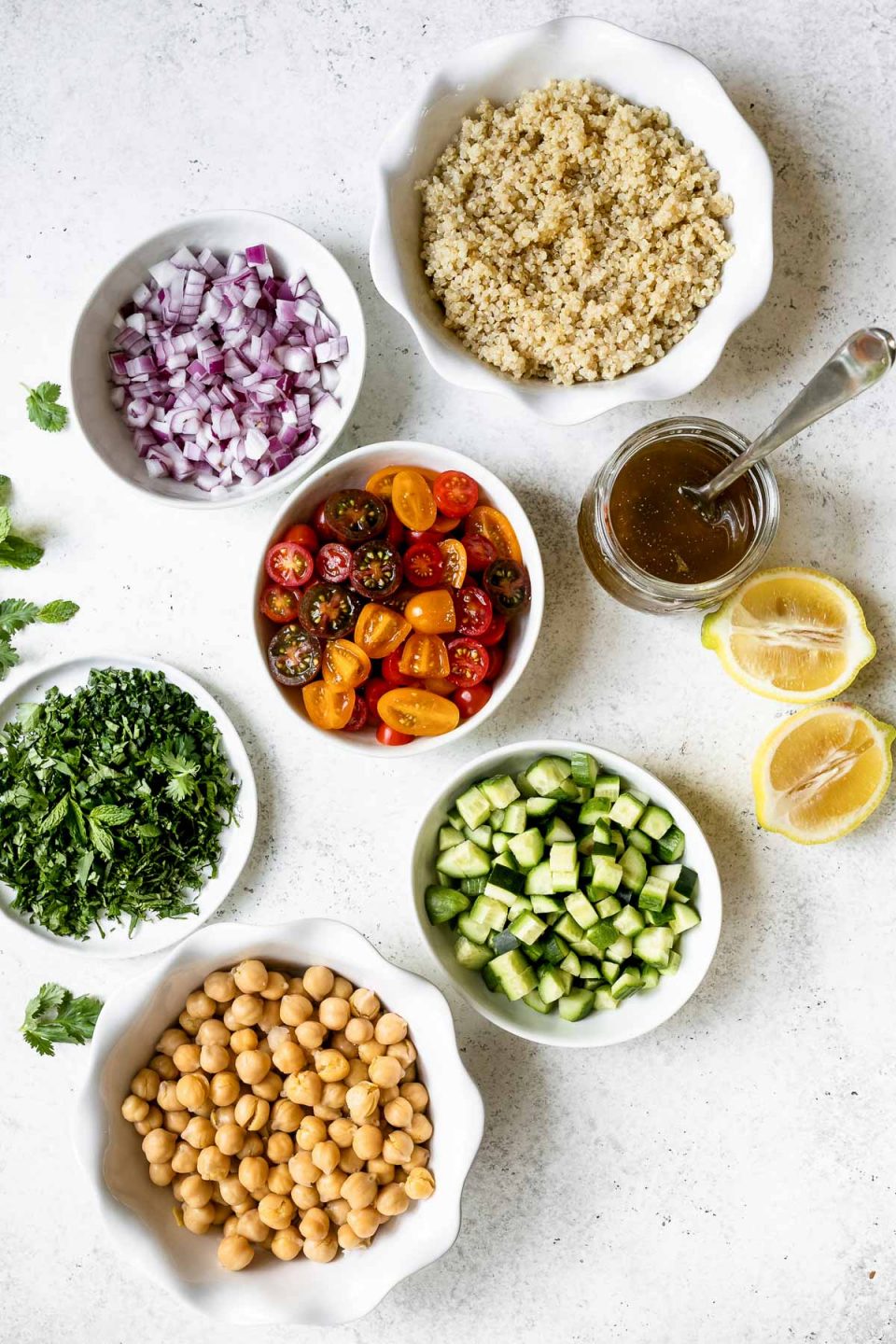 Quinoa salad ingredients in individual small bowls, arranged on a white surface - red onion, quinoa, tomatoes, salad dressing, lemon, cucumber, chickpeas & fresh herbs.