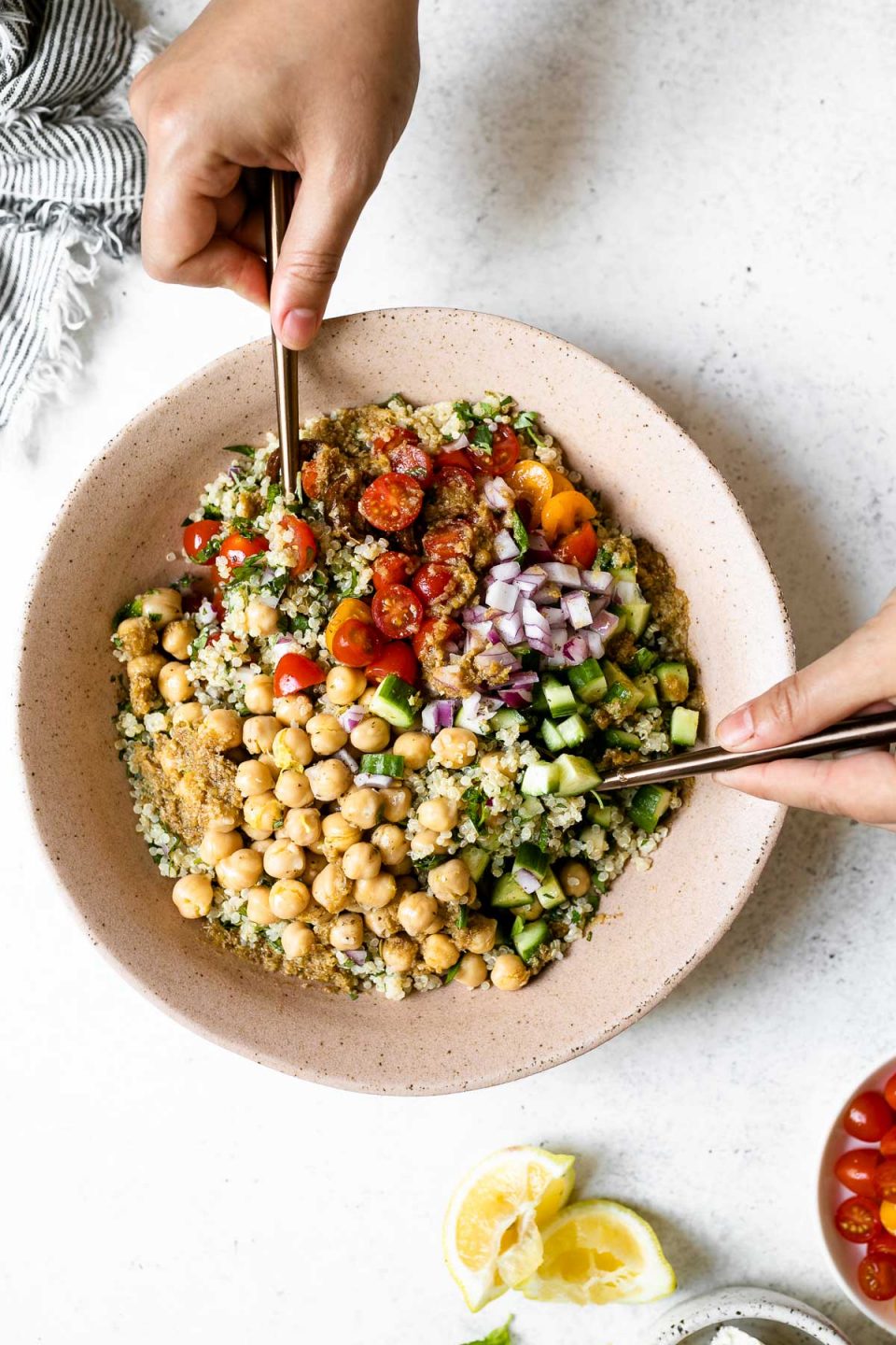 Falafel-ish Quinoa Salad in a large, pink serving bowl. A woman's hands shown tossing the salad with copper serving utensils. The salad bowl is placed on a white surface, & surrounding it are a linen napkin, & some lemon wedges.