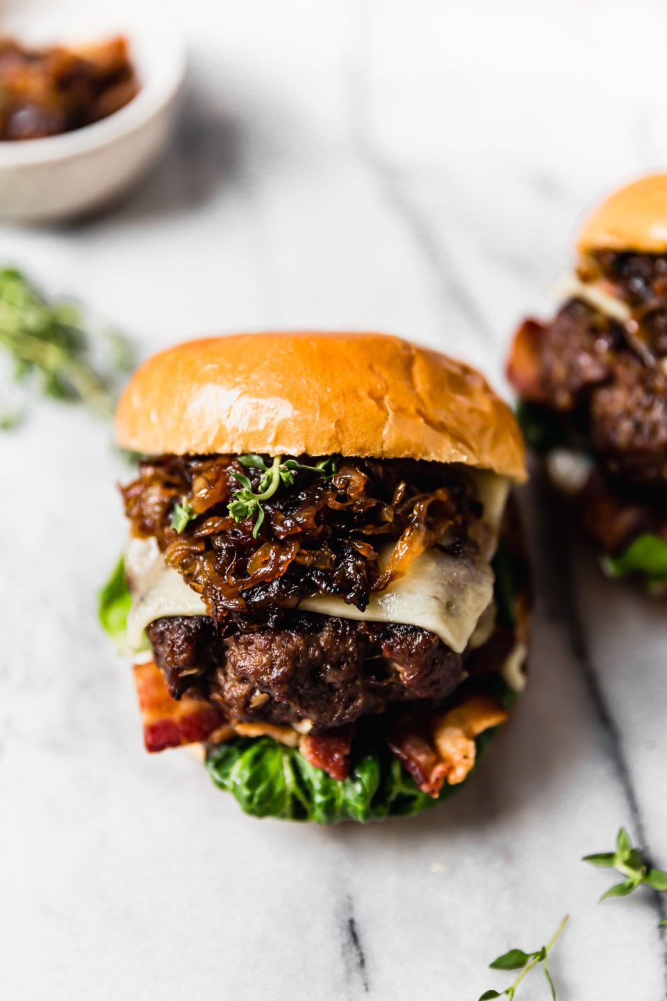 2 French onion burgers on a white marble surface. The burgers are fixed with lettuce, bacon, cheese & caramelized onions, & served on brioche burger buns.