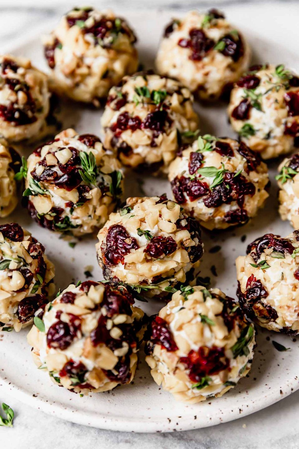 Herbed goat cheese balls, coated in chopped nuts & tart cherries, on a small, speckled white plate.