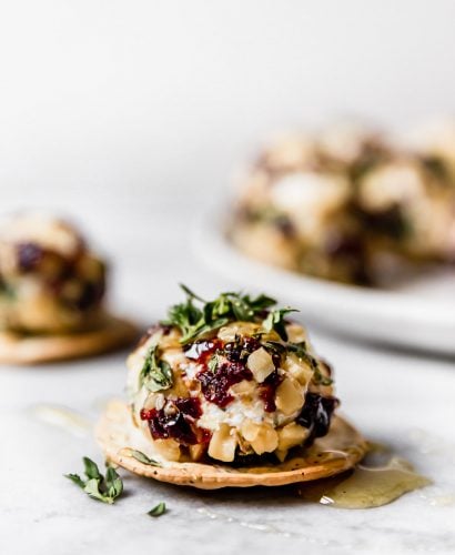 Goat cheese truffle, coated with chopped nuts, tart cherries, & fresh herbs, served on a cracker, topped with honey. There are more goat cheese balls in the background on a small white plate.