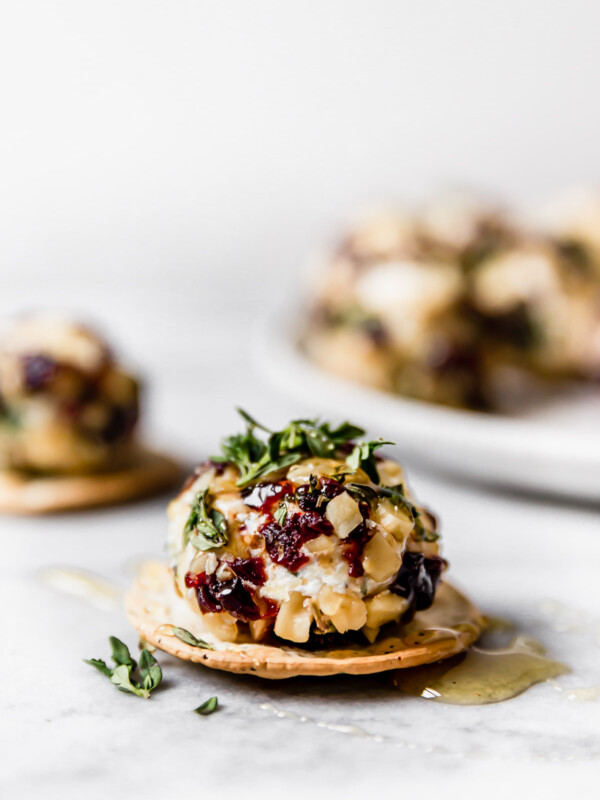 Goat cheese truffle, coated with chopped nuts, tart cherries, & fresh herbs, served on a cracker, topped with honey. There are more goat cheese balls in the background on a small white plate.