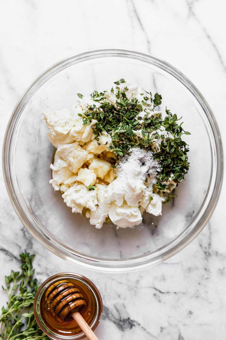 Goat cheese, honey, & fresh thyme in a glass mixing bowl, on a white marble surface next to fresh thyme & a small jar of honey.